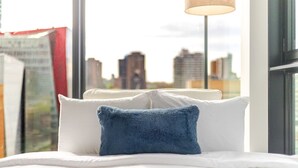 En säng med vita sängkläder och en blå kudde, ett fönster med utsikt över stadens byggnader och en lampa på ett stativ.
