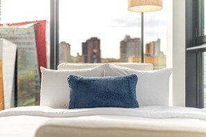 A bed with white bedding and a blue pillow, a window with a view of city buildings, and a lamp on a stand. - Luxe Central Retreat | Free Parking & Pool (Montréal)