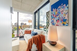 A modern living room with a large window, a white sofa, a lamp, and a colorful abstract painting. - Luxe Central Retreat | Free Parking & Pool (Montréal)