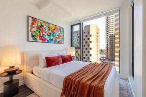 A modern hotel room with a large bed, two bedside tables, a colorful abstract painting, and a view of tall buildings. - Luxury Stay | Free Parking (Montréal)