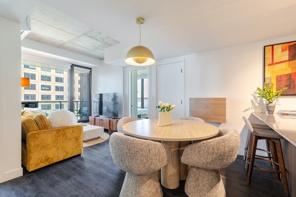 Een moderne woonkamer met een ronde eettafel, zachte zitplaatsen, een televisie en grote ramen met uitzicht op de stad.