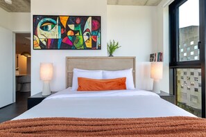 A modern hotel room with a large bed, a colorful abstract painting, a view through the window, and a small plant on the nightstand. - City Haven | Free Parking (Montréal)