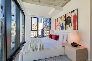 A modern hotel room with a large window, a bed with white bedding, a red pillow, and abstract art on the wall. - Modern Montreal Escape | Pools Gym Cinema Balcony (Montreal)