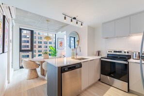 A modern kitchen with a dishwasher, oven, and a large island. There is a dining area with a table and chairs. - Urban Prestige Apartment | Free Parking & Pool (Montréal)