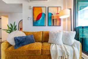 A mustard-colored sofa with fluffy pillows, a white throw blanket, and abstract art on the wall. - Modern Escape | Free Parking & Rooftop Pool (Montréal)