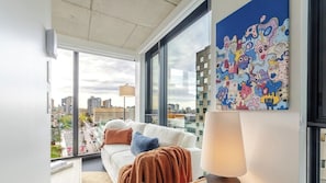 A modern living room with a large window, a white sofa, a colorful abstract painting, and a lamp. - Urban Oasis Balcony Stay | Free Parking & Pool (Montréal)