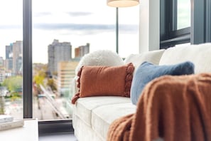 Ein Sofa mit mehreren Kissen, ein Fenster mit Blick auf eine Stadtlandschaft und eine Lampe.