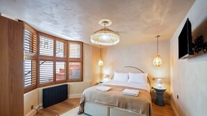 Een slaapkamer met een bed, houten jaloezieën, een radiator, een hanglamp en een aan de muur bevestigde televisie.