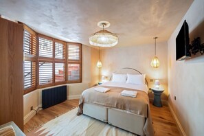Ein Schlafzimmer mit Bett, Holzjalousien, Heizkörper, Pendelleuchte und an der Wand befestigtem Fernseher.