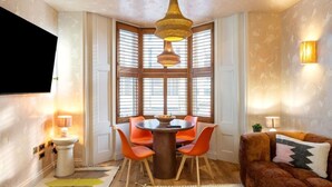Een eethoek met een ronde tafel, oranje stoelen, een hanglamp en een erker met jaloezieën.