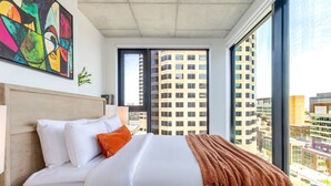 Een moderne hotelkamer met een groot bed, uitzicht op de stad en een kleurrijk abstract schilderij aan de muur.