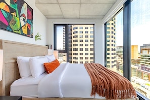 Ein modernes Hotelzimmer mit einem großen Bett, Blick auf die Stadt und einem farbenfrohen abstrakten Gemälde an der Wand.