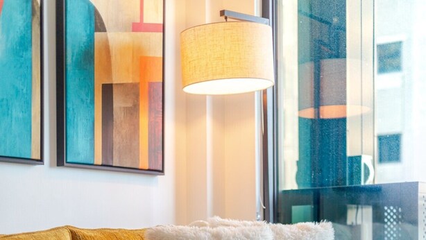 Een gele bank met zachte kussens, een witte plaid en een houten salontafel in een moderne woonkamer met abstracte kunst aan de muur en een groot raam.