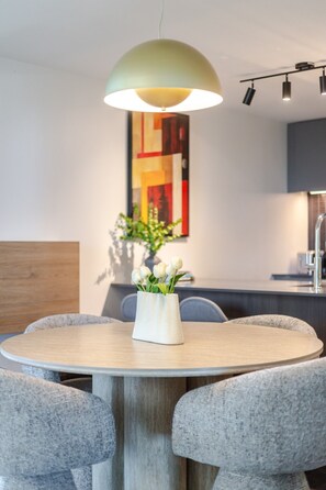 Une table de salle à manger ronde, sur laquelle est posé un vase blanc rempli de fleurs, des chaises à revêtement gris, un luminaire suspendu et, au mur, un tableau abstrait moderne.