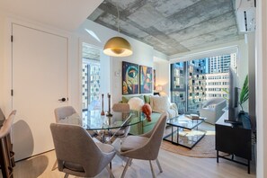 A modern living room with a glass coffee table, a green sofa, and a large window offering a city view. - Executive Apartment | Free Parking & Pool (Montreal)
