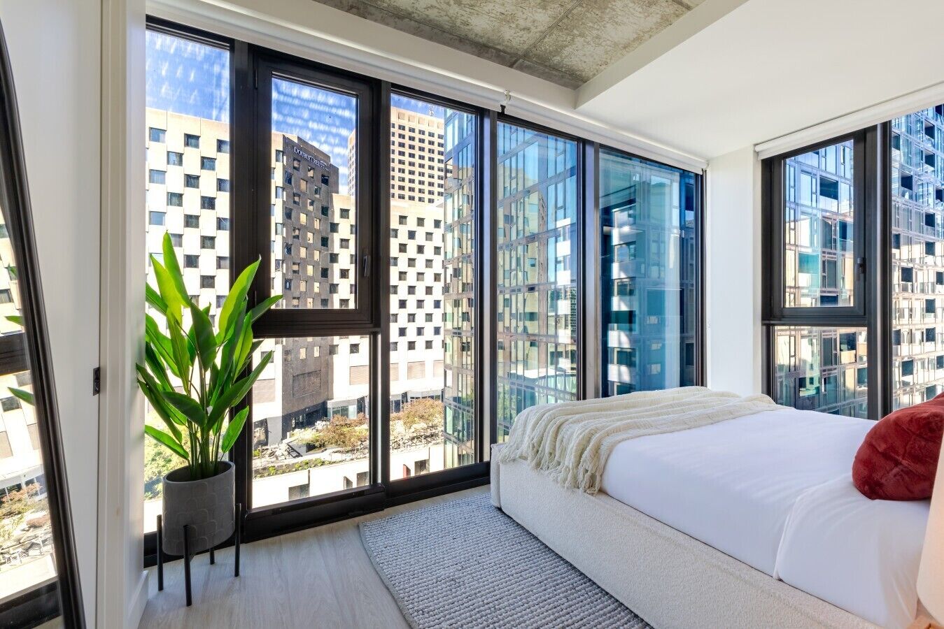 Un dormitorio moderno con ventanas amplias, una cama, una planta y vistas a edificios de la ciudad.