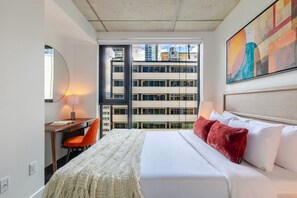 Hotellihuone, jossa on sänky, työpöytä, tuoli, suuri ikkuna, josta on näkymä rakennukseen, ja taulu seinällä.
