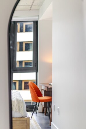 Ein Zimmer mit einem Bett, einem orangefarbenen Sessel, einem Schreibtisch und einem Fenster mit Blick auf Gebäude.