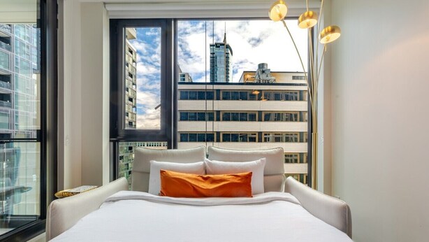 Une chambre d’hôtel moderne avec un grand lit, une vue sur le paysage urbain grâce à une grande fenêtre et un élégant luminaire suspendu.