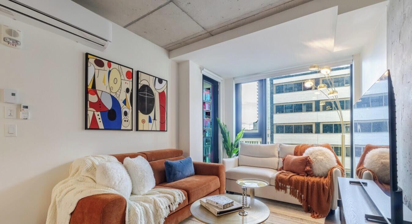 Un salon moderne avec un canapé, un fauteuil et une table basse. Les murs sont ornés de tableaux abstraits et une grande fenêtre offre une vue sur les bâtiments.