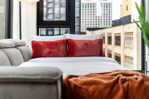 A modern bedroom with a bed, grey sofa, and a view of city buildings. - Downtown Comfort Apartment | Free Parking & Pool (Montréal)