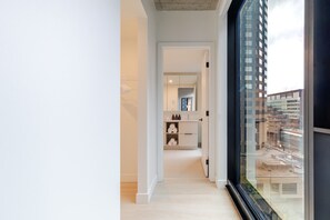 Un intérieur moderne avec un couloir menant à une salle de bain, une grande fenêtre offrant une vue sur la ville et un design minimaliste.