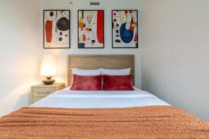 Une chambre d’hôtel avec un lit, une table de chevet avec une lampe et trois tableaux abstraits accrochés au mur.