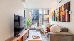 Ein modernes Wohnzimmer mit Fernseher, Couch, Kissen und einem Holztisch, mit großem Fenster und Blick auf die Stadt.