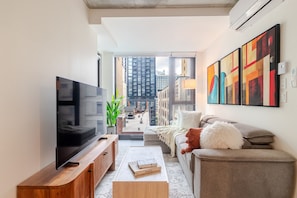 Ein modernes Wohnzimmer mit Fernseher, Couch, Kissen und einem Holztisch, mit großem Fenster und Blick auf die Stadt.