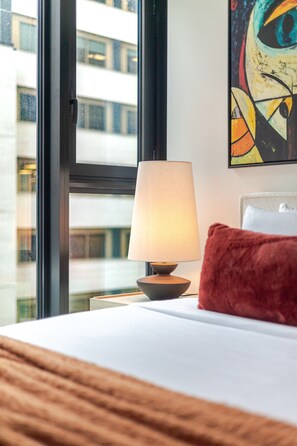 Una habitació d'hotel amb un llit, una làmpada, un coixí vermell i una finestra que mostra edificis.