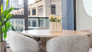 Ein runder Tisch mit einer Vase weißer Blumen, grauen Polsterstühlen und Blick auf Gebäude durch große Fenster.