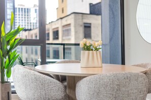 Ein runder Tisch mit einer Vase weißer Blumen, grauen Polsterstühlen und Blick auf Gebäude durch große Fenster.