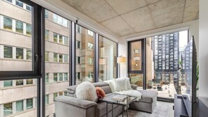 Uma sala de estar moderna com um sofá, uma mesa de centro e um abajur. Janelas amplas oferecem vista para o horizonte da cidade.