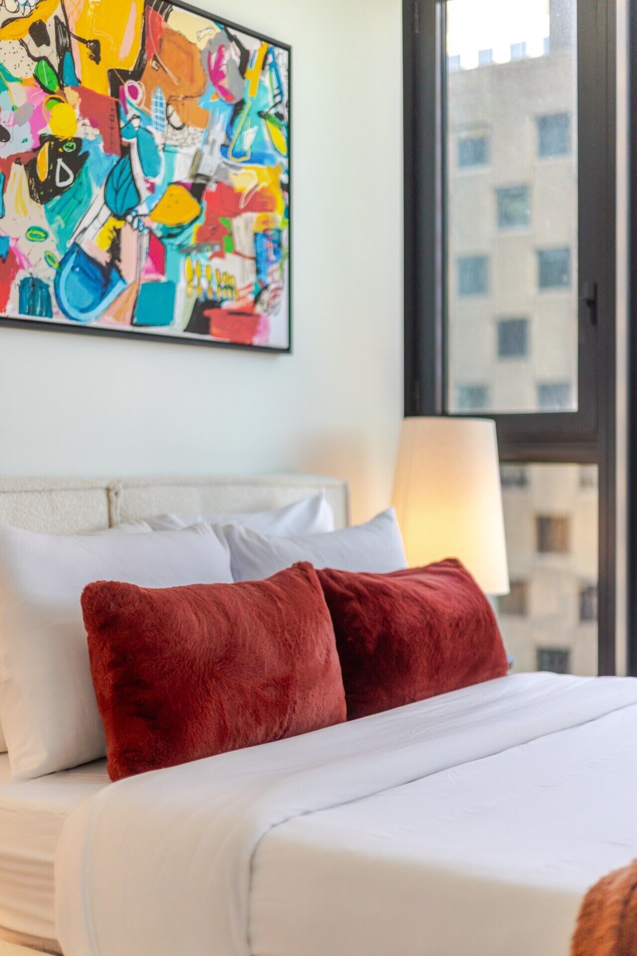 A hotel room with a bed, pillows, a window, and a colorful abstract painting on the wall.