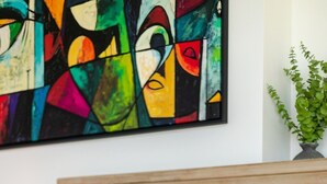 Een bed met witte en oranje beddengoed, een houten hoofdbord en een kleurrijk abstract schilderij aan de muur.