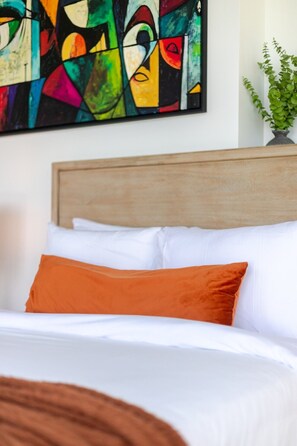 Ein Bett mit weißer und orangefarbener Bettwäsche, ein hölzernes Kopfteil und ein farbenfrohes abstraktes Gemälde an der Wand.
