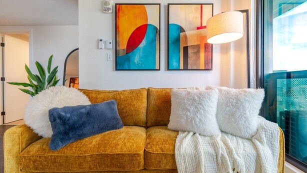 Een mosterdgele bank met pluizige kussens, een witte plaid en abstracte kunst aan de muur.