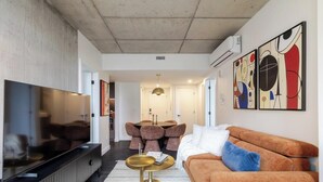 A modern living room with a flat-screen TV, a sofa, a dining area with chairs, and abstract wall art. - Golden Stay | Free Parking & Pool (Montreal)