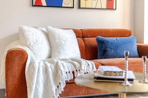 A living room with a sofa, two pillows, a white throw blanket, and a coffee table with books and a decorative item. - Golden Stay | Free Parking & Pool (Montreal)