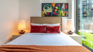 Ein Hotelzimmer mit einem Bett, zwei Nachttischlampen, einem farbenfrohen abstrakten Gemälde über dem Bett und einem großen Fenster mit Blick auf Grünflächen.