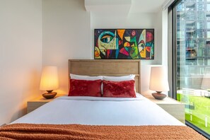 A hotel room with a bed, two bedside lamps, a colorful abstract painting above the bed, and a large window with a view of greenery. - Montreal Skyline Escape | Free Parking & Pool (Montreal)