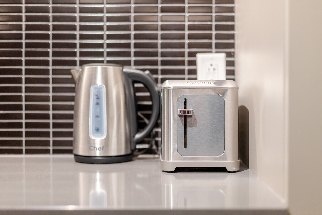 Ein Edelstahlkessel und ein Toaster stehen auf einer Arbeitsplatte. Im Hintergrund ist ein Fenster mit Jalousien zu sehen.