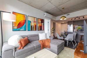 A modern living room with a grey sofa, a coffee table, and a dining area with chairs. There are abstract paintings on the wall, a pendant light, and an air conditioning unit. - Downtown Poolside Haven | Free Parking & Pool (Montreal)