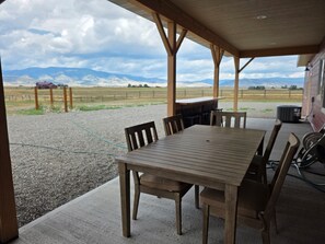 An outdoor covered patio with a wooden table and chairs, a gravel ground, and a view of a rural landscape with mountains in the distance. - Bear Claw Mountain House (Livingston)