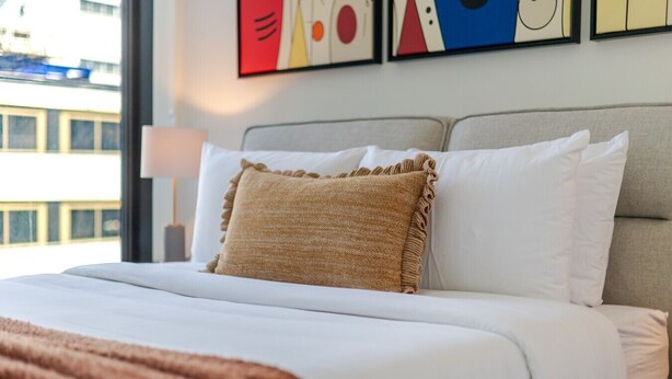 Un letto rifatto con lenzuola bianche e una coperta marrone, una testiera grigia e un muro con arte astratta.