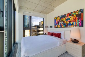 Ein modernes Hotelzimmer mit einem großen Bett, einem farbenfrohen abstrakten Gemälde und Blick auf eine Stadtlandschaft durch ein großes Fenster.