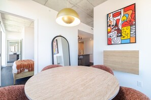 A modern room with a round table, a bed with a brown blanket, a kitchen area, and a colorful abstract painting on the wall. - Luxury Urban Retreat | Pools Gym Sauna & Balcony (Montreal)