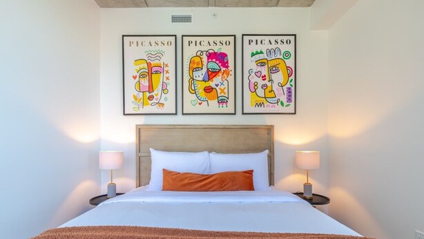 Une chambre d’hôtel avec un lit, deux lampes de chevet, un plaid marron et trois œuvres d’art de Picasso accrochées au mur.