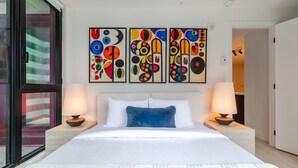 Quarto de hotel com uma cama grande, duas mesas de cabeceira, uma janela e três pinturas abstratas na parede.