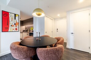 A modern dining area with a round table, four upholstered chairs, a pendant light, and a kitchen in the background. - Modern City Retreat | Balcony + Resort Amenities (Montreal)
